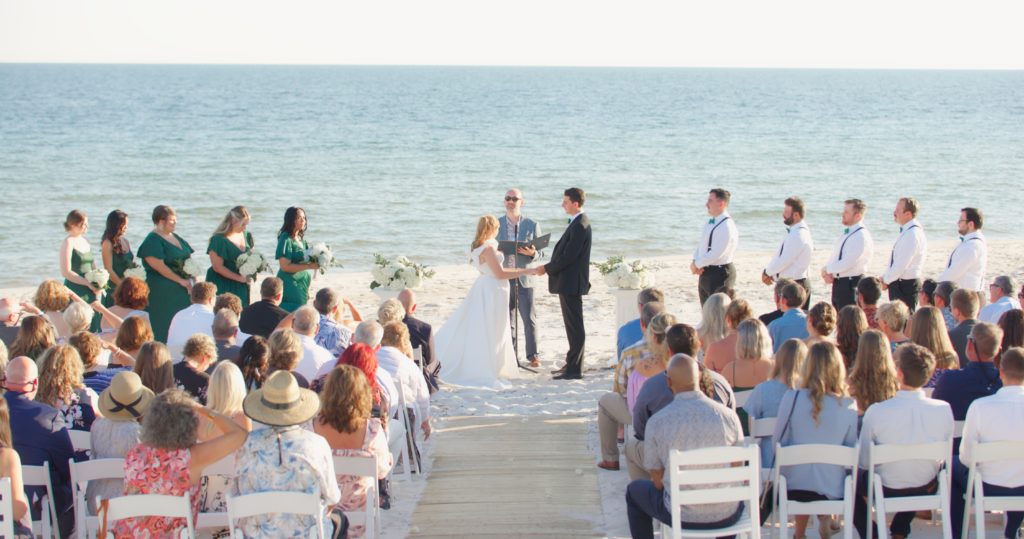 During the wedding ceremony section of our wedding videography timeline, a couple gets married on the beach with their wedding party on either side of them. Their guests watch.